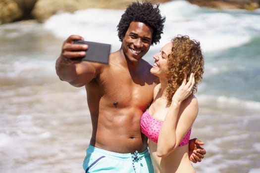 Smiling multiracial couple embracing on beach near waving sea while taking selfie on smartphone during summer vacation