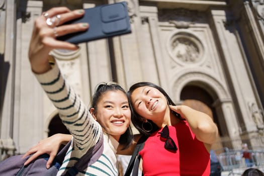 Positive young Asian female tourists in casual clothes with backpacks smiling and taking selfie on smartphone against Cathedral of Granada during trip through Spain