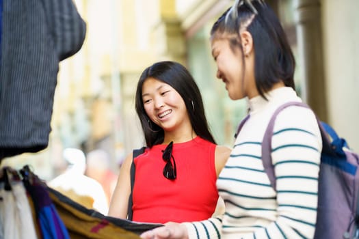 Asian girls, in casual clothes with backpack smiling while shopping clothes on street market in city during vacation