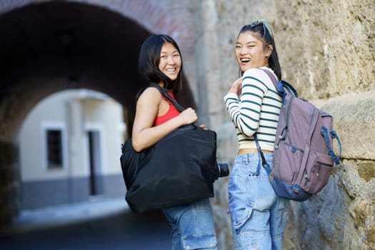 Cheerful Asian women tourists with belongings looking over shoulder at camera with backpack while standing near stone wall of building