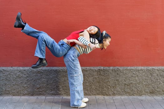 Side view of cheerful Asian woman in casual clothes lifting smiling friend on back while standing against red wall
