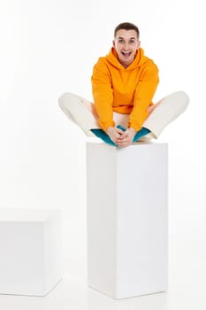 Full length photo of young man in orange sweatshirt sitting on white cube on white background.
