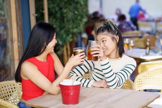 Positive young Asian female friends in casual clothes sitting together at wooden table, and clinking glasses of fresh beverages while looking at each other during meeting in cafe