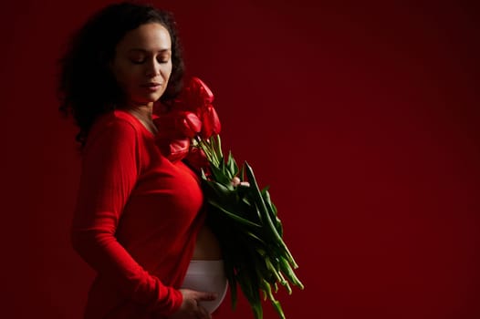 Confident sensual delightful stunning beautiful pregnant woman in red wear, touching her belly, posing with a bunch of red tulips on isolated color background with copy ad space. Pregnancy. Maternity