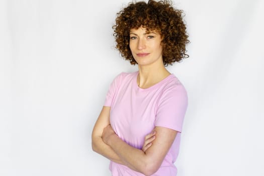 Adult curly haired woman in casual clothes standing with crossed hands and looking at camera while smiling in light studio