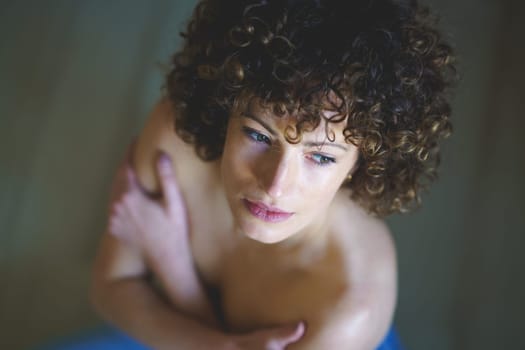 High angle of woman with curly hair, with her bare shoulders gazing through the window with her gaze lost while sitting on floor with crossed arms