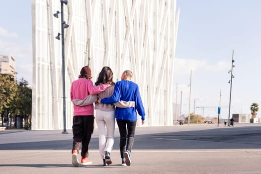 rear view of three unrecognizable friends, one man and two women, embracing while walking on a sunny day, concept of friendship and urban lifestyle