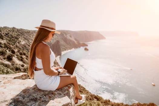 Successful business woman in yellow hat working on laptop by the sea. Pretty lady typing on computer at summer day outdoors. Freelance, travel and holidays concept.