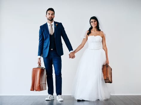 We have our travel shoes on. Studio shot of a newly married couple holding hand and carrying bags while standing against a gray background