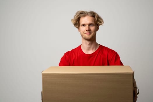 Delivery service. Young smiling courier holding cardboard box. Happy young delivery man in red t-shirt standing with parcel isolated on white background