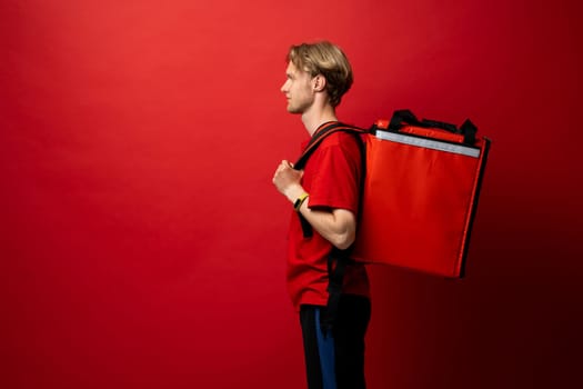 Young courier with thermal bag on red background, space for text. Food delivery service. Delivery guy in a red t-shirt uniform work as courier and holds red thermal food backpack. Service concept