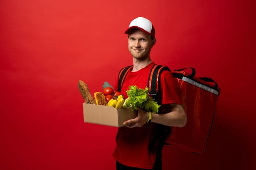 Delivery male employee in a red uniform holds paper cardboard package with organic groceries, fruits on a red background. Products delivery from shop or restaurant to home. Courier service