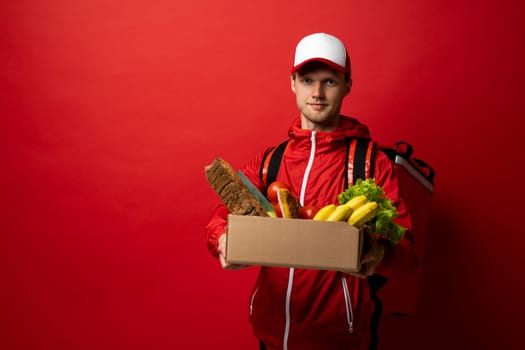 Delivery man in a red uniform holding paper box full of groceries. Online shopping and express delivery. Quality service of a restaurant