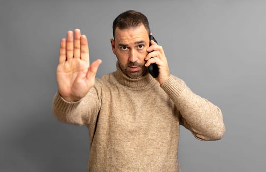 Bearded Latino man in his 40s wearing a beige turtleneck talking on the cellphone with a serious expression while raising his hand in a stop sign, isolated on yellow studio background.