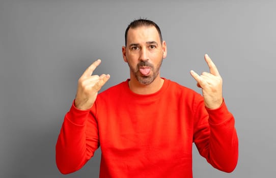 Latino man with a beard in his 40s wearing a red sweater making the rock gesture while sticking out his tongue in a crazy attitude, isolated on yellow studio background