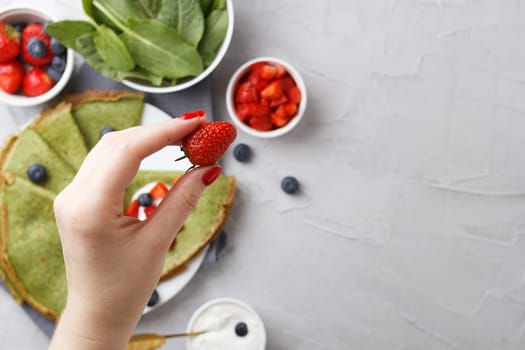 A woman's hand holds strawberries over a plate with spinach pancakes with berries and sour cream on a gray background with textiles. copy space.