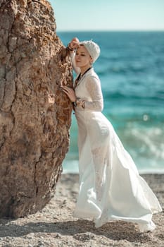 Woman white dress sea stones rocks.Middle-aged woman looks good with blond hair, boho style in a white long dress on beach jewelry around her neck and arms