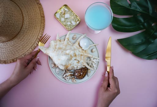 Creative photography. The concept of summer vacation memories. In the women's hands are golden eating utensils, on the table is a straw hat, a sheet of monstera and there is a plate with seashells.