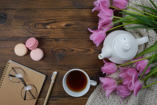 On a wooden table there is a white teapot, a cup of tea, a notebook with a frame of glasses, and beautiful tulips.