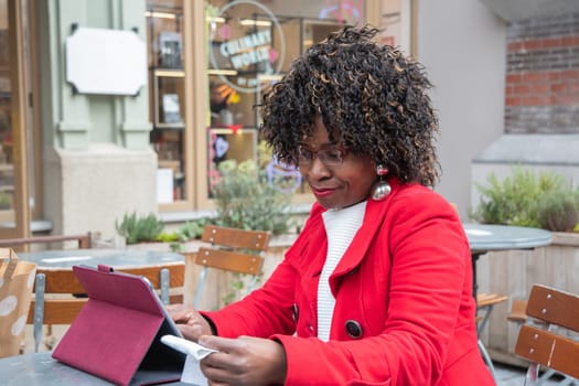 a young african american woman in a red coat sits at a table in a cafe and checking bill after shopping using a tablet, financial problems crisis, proper planning of expenses, High quality photo