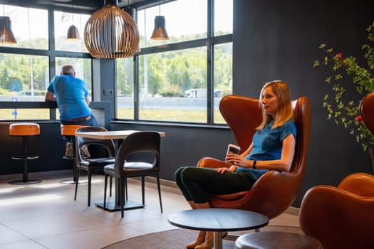 millennial blonde businesswoman sits at a table in a cafe and drinks coffee from a cardboard cup. High quality photo