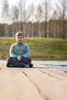 A boy is sitting on a bridge in a green park. The path is a bridge over the lake. Long wooden flooring