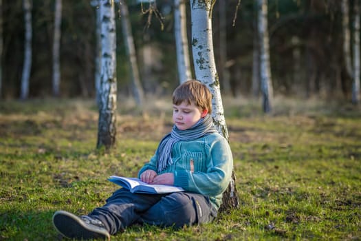 A handsome boy in a scarf is in a spring park, sitting under a birch tree, reading a book and enjoying his dreams. A happy child. Portrait