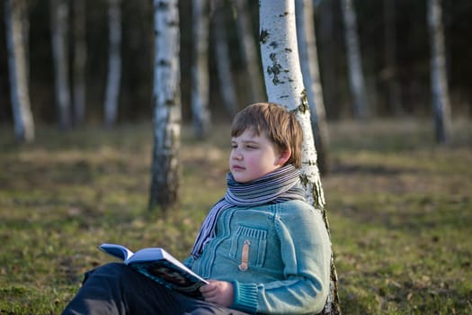 A handsome boy in a scarf is in a spring park, sitting under a birch tree, reading a book and enjoying his dreams. A happy child. Portrait