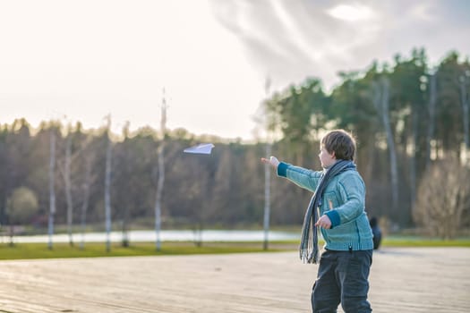 A handsome boy in a scarf is in a spring park, happily launching a paper airplane. A happy child. Portrait