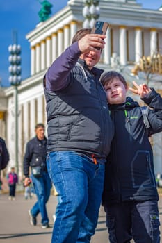 2023-04-08. Moscow, Russia. A man and a boy take a selfie on a smartphone in the city center near an architectural building. Moscow, VDNH.