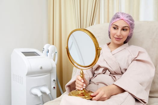 Happy young woman sitting in medical chair and looking in the mirror. She is satisfied after successful beauty treatment with hyaluronic acid fillers