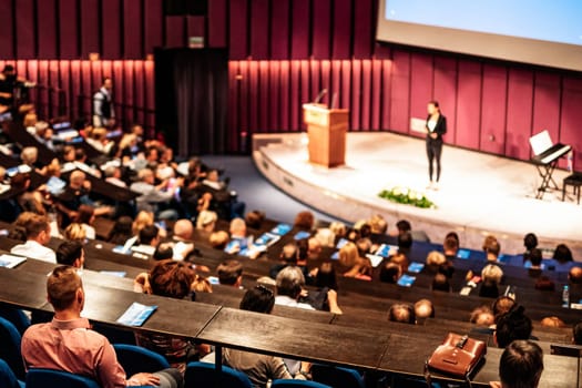 Business and entrepreneurship symposium. Female speaker giving a talk at business meeting. Audience in conference hall. Rear view of unrecognized participant in audience.