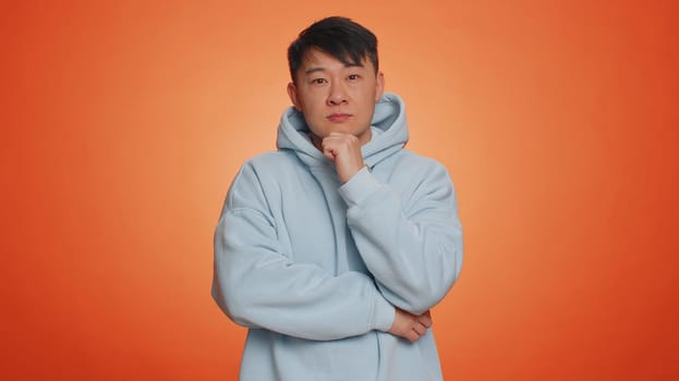 Thoughtful clever asian handsome man rubbing his chin and looking aside with pensive expression, pondering a solution, doubting question. Adult guy on orange studio background. People lifestyles