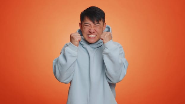 Asian man in hoodie screams yell from stress tension problems feels horror hopelessness fear panic surprise shock expresses gestures rage. Handsome chinese guy isolated on orange studio background