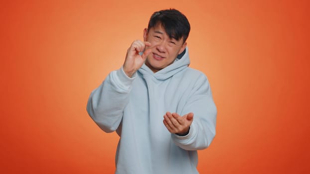 Need some more, please give me. Asian man showing a little bit gesture with sceptic smile, showing minimum sign, measuring small size. Handsome chinese guy isolated alone on orange studio background
