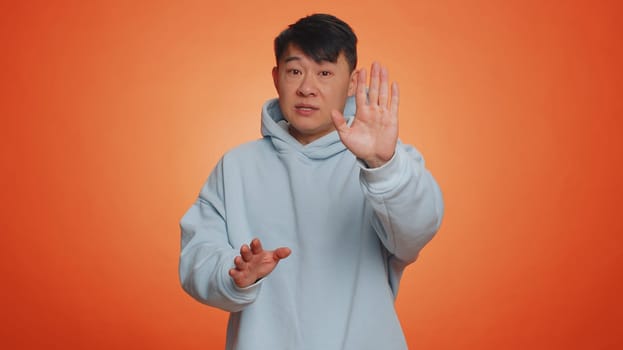 Asian man say No hold palm folded crossed hands in stop gesture, warning of finish, prohibited access, declining communication, body language, danger. Adult guy on orange background. People lifestyles