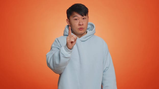 Asian handsome man shakes finger and saying No, be careful, scolding and giving advice to avoid danger mistake, disapproval sign. Adult guy isolated alone on orange studio background. People lifestyle