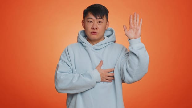 I swear to be honest. Sincere responsible asian handsome man raising hand to take oath, promising to be honest and to tell truth, keeping hand on chest. Adult guy isolated on orange studio background