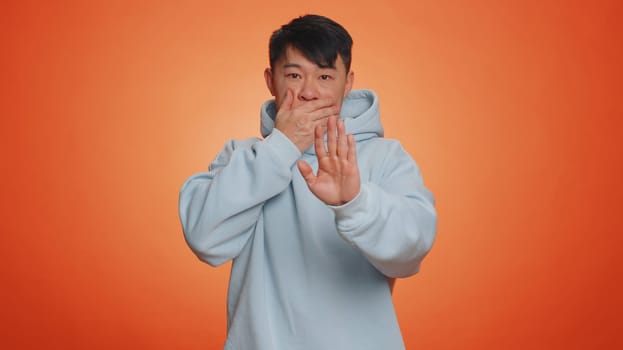 I will not say anyone. Frightened asian man closing mouth with hands, looking intimidated scared at camera, gestures no, refusing to tell terrible secret, unbelievable truth. Guy on orange background