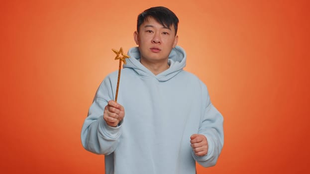 Magician wizard man gesturing with magic wand fairy stick, making wish come true, casting magician spell, advertising holidays sale discount. Handsome asian guy isolated on orange studio background
