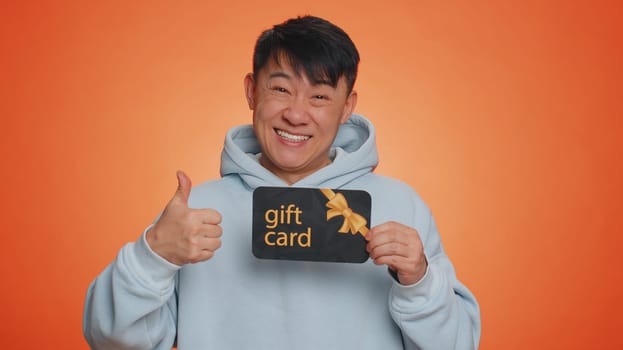 Handsome chinese man showing pointing on gift discount certificate voucher coupon for store holidays sale. Asian guy with shopping christmas surprise card isolated alone on orange studio background