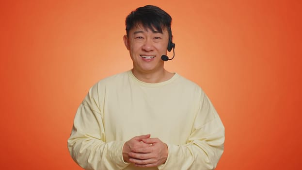 Smiling asian man wearing headset, freelance worker, call center or support service operator helpline, having talk with client or colleague, communication support. Handsome guy on orange background
