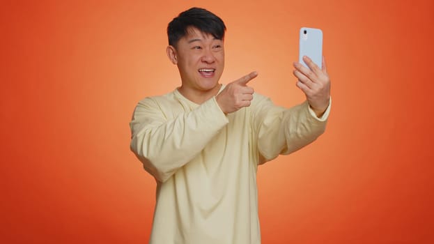 Asian man traveler blogger, taking selfie on smartphone, communicating video call online with family or friends, live translation stream. Handsome chinese guy isolated on orange background, indoors