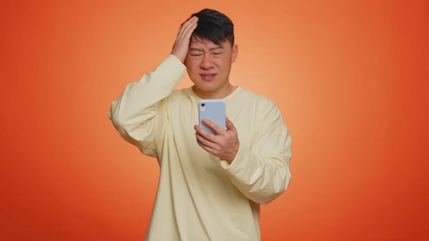 Asian man use mobile cell phone typing browsing, loses becoming surprised sudden lottery results, bad fortune, loss, fail. Handsome chinese guy isolated alone on orange studio wall background, indoors