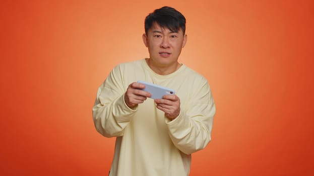 Worried funny addicted asian man enthusiastically playing racing video games on mobile phone. Handsome adult guy using smartphone gadget app with drive simulator isolated on orange studio background