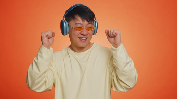 Happy asian man in headphones listening music and dancing disco fooling around having fun expressive gesticulating hands, relaxing on party. Chinese guy isolated alone on studio orange wall background