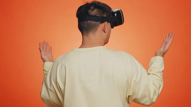 Man using headset helmet to play simulation game app online. Watching virtual reality 3D 360 video content. Guy in VR goggles isolated on studio orange background. Futuristic technology. Rear view