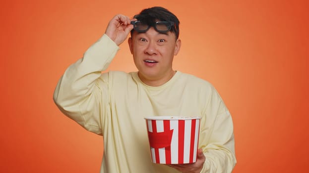 Excited asian man in 3D glasses eating popcorn and watching interesting tv serial, sport game, film, online social media movie content. Happy guy enjoying domestic entertainment on orange background