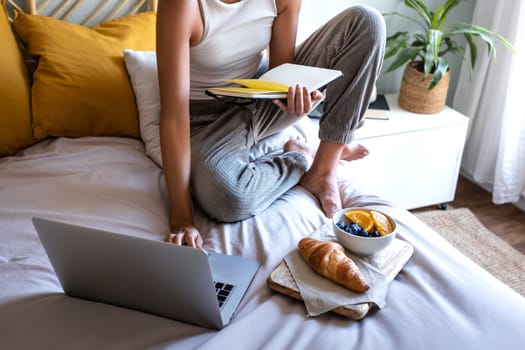 Unrecognizable woman eating breakfast in bed relaxing at home while working with laptop. Lifestyle concept.