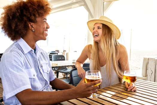Young multiracial couple on vacation enjoying time together, laughing at a beach bar drinking beer. Lifestyle and summertime concept.